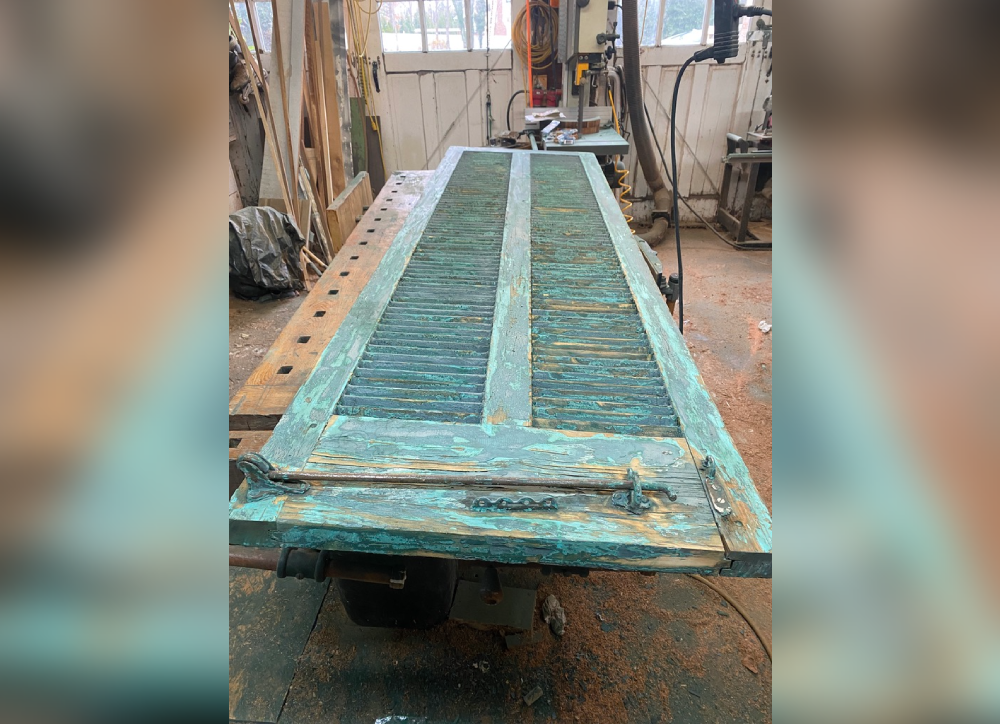 Weathered wooden shutters with a turquoise finish, resting on a workbench in a workshop setting.