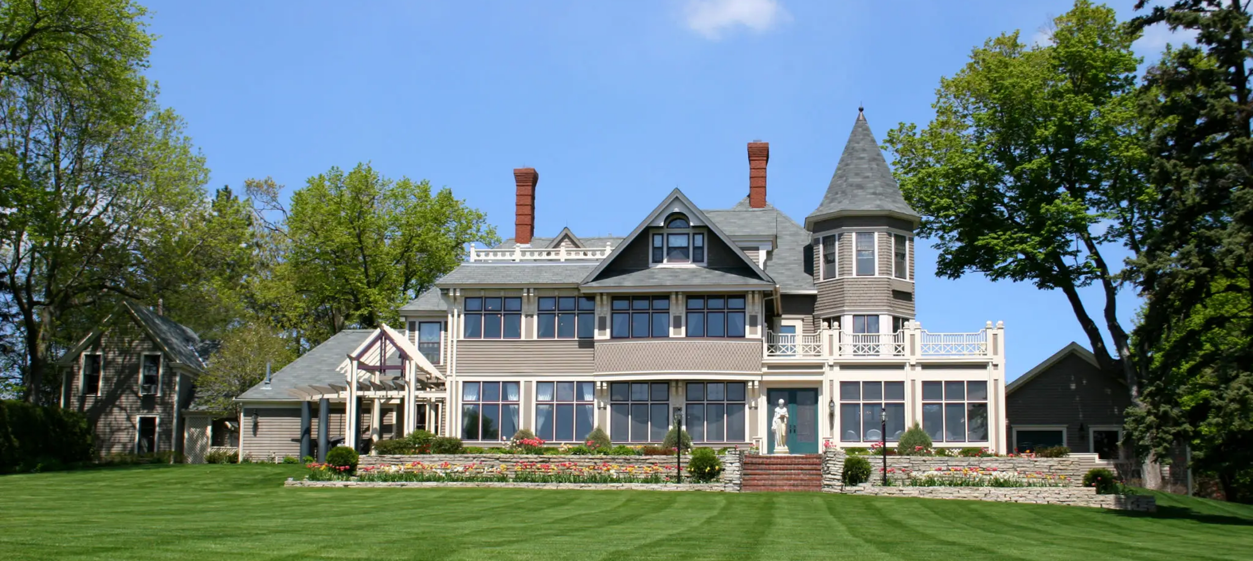 Victorian-style mansion with a turret, surrounded by lush greenery and a well-maintained lawn, showcasing architectural elegance.